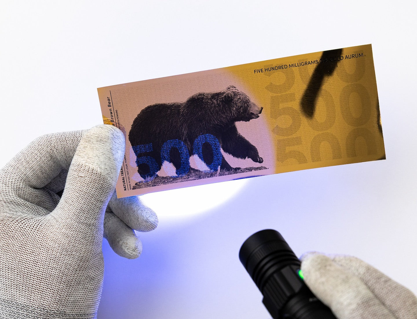a blacklight pointed towards the left of a half gram gold aurum bill revealing the hidden UV design highlighting a 500 to the left of the bill