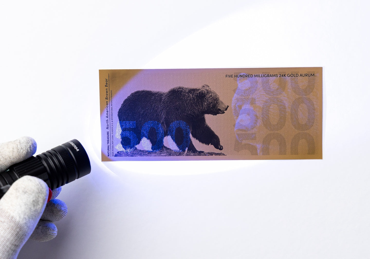 a blacklight held to the left of a half gram gold aurum bill revealing the hidden UV design highlighting a 500 to the left of the bill