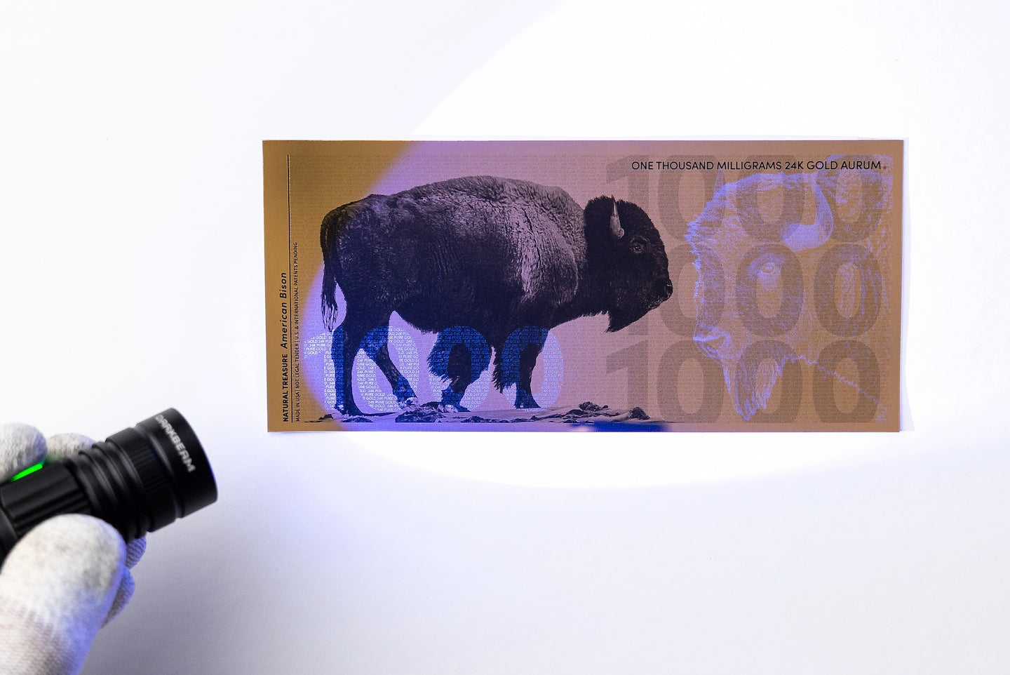 a blacklight held to the left of a gold aurum bill revealing the hidden UV design highlighting a 1000 to the left of the bill