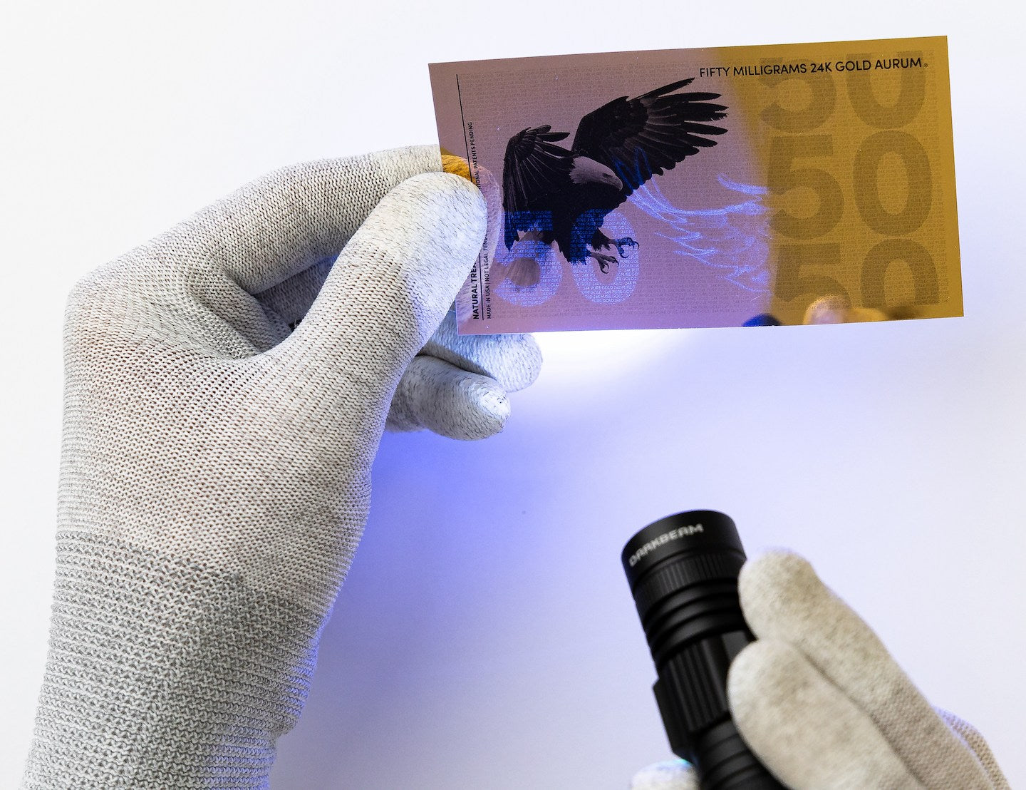 a black light pointed towards the left of a 50mg gold aurum bill revealing the hidden UV design highlighting the number 50