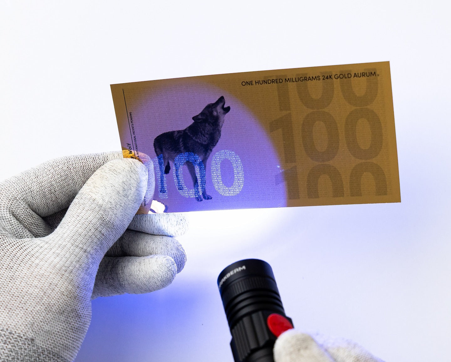 a blacklight pointed towards the left of a 100mg gold aurum bill revealing the hidden UV features highlighting the number 100